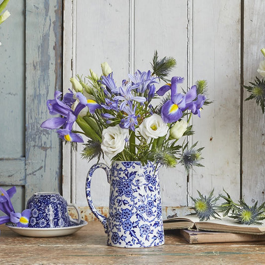 A styled tabletop arrangement of blue-and-white ceramic pitchers, cups, and a vase holding white roses and purple flowers, set against a rustic light blue wooden wall.