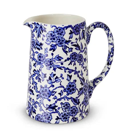 A white ceramic pitcher decorated with a blue floral and vine pattern, featuring a spout and a curved handle, set against a plain white background.