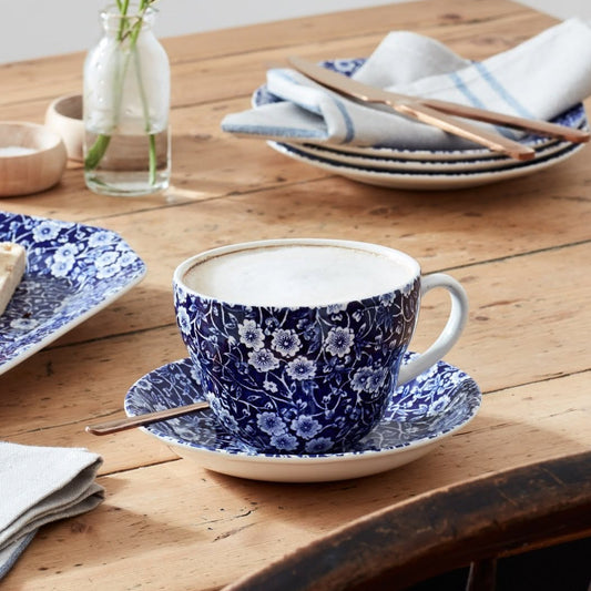 A blue and white Burleigh calico floral ceramic teacup and saucer set on a rustic wooden table, styled with a matching plate of biscotti, folded striped napkins, and a small glass vase.