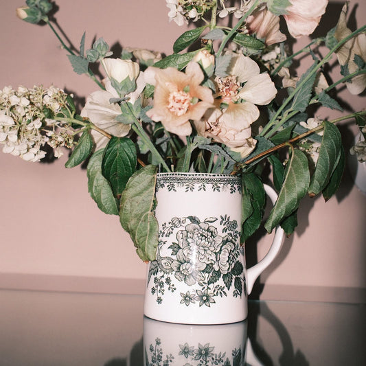 A white ceramic pitcher with a green floral print, filled with a bouquet of pale pink and cream flowers with green leaves, set against a soft pink background.